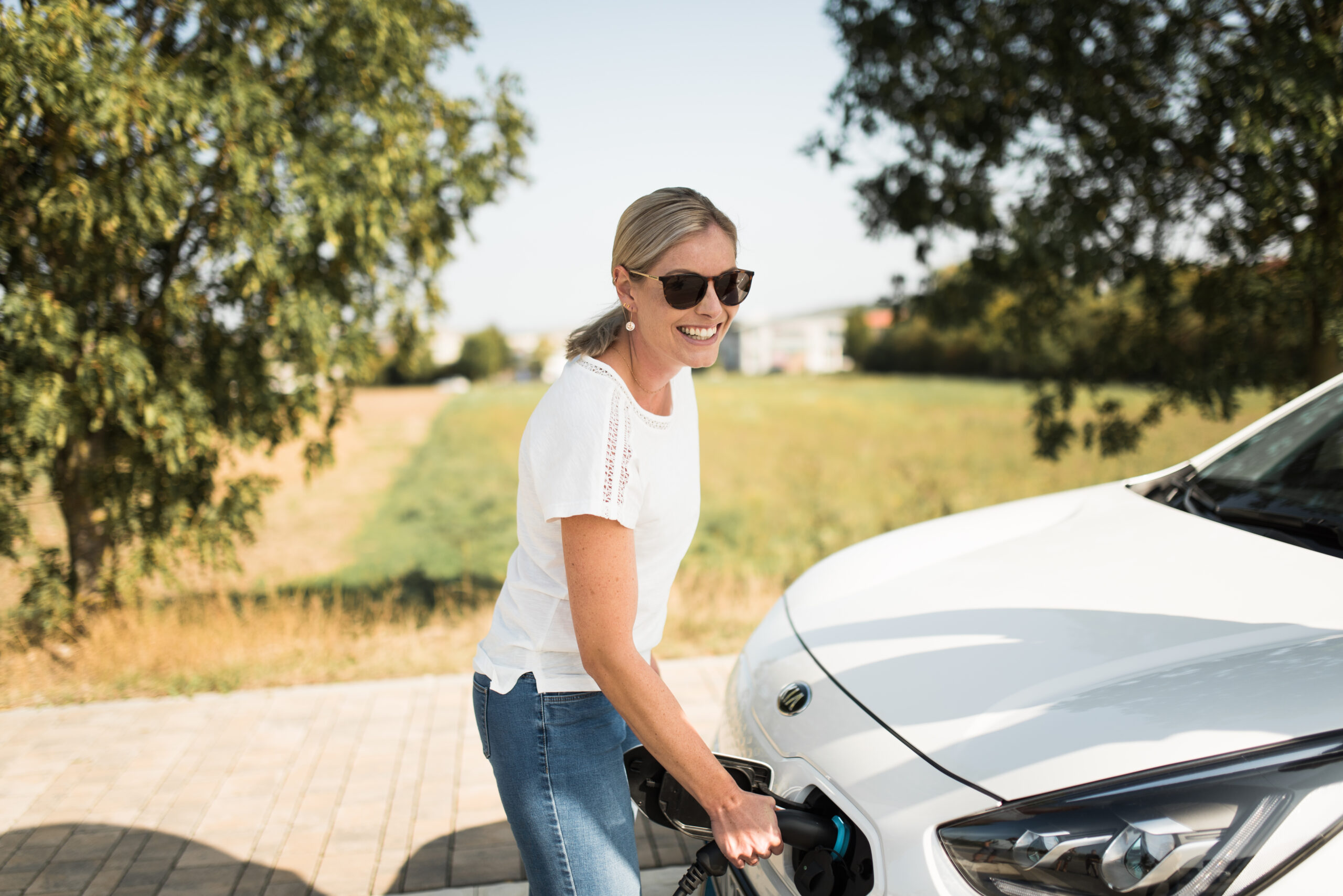 Frau mit Sonnenbrille läd lachend weißes E-Auto bei gutem Wetter