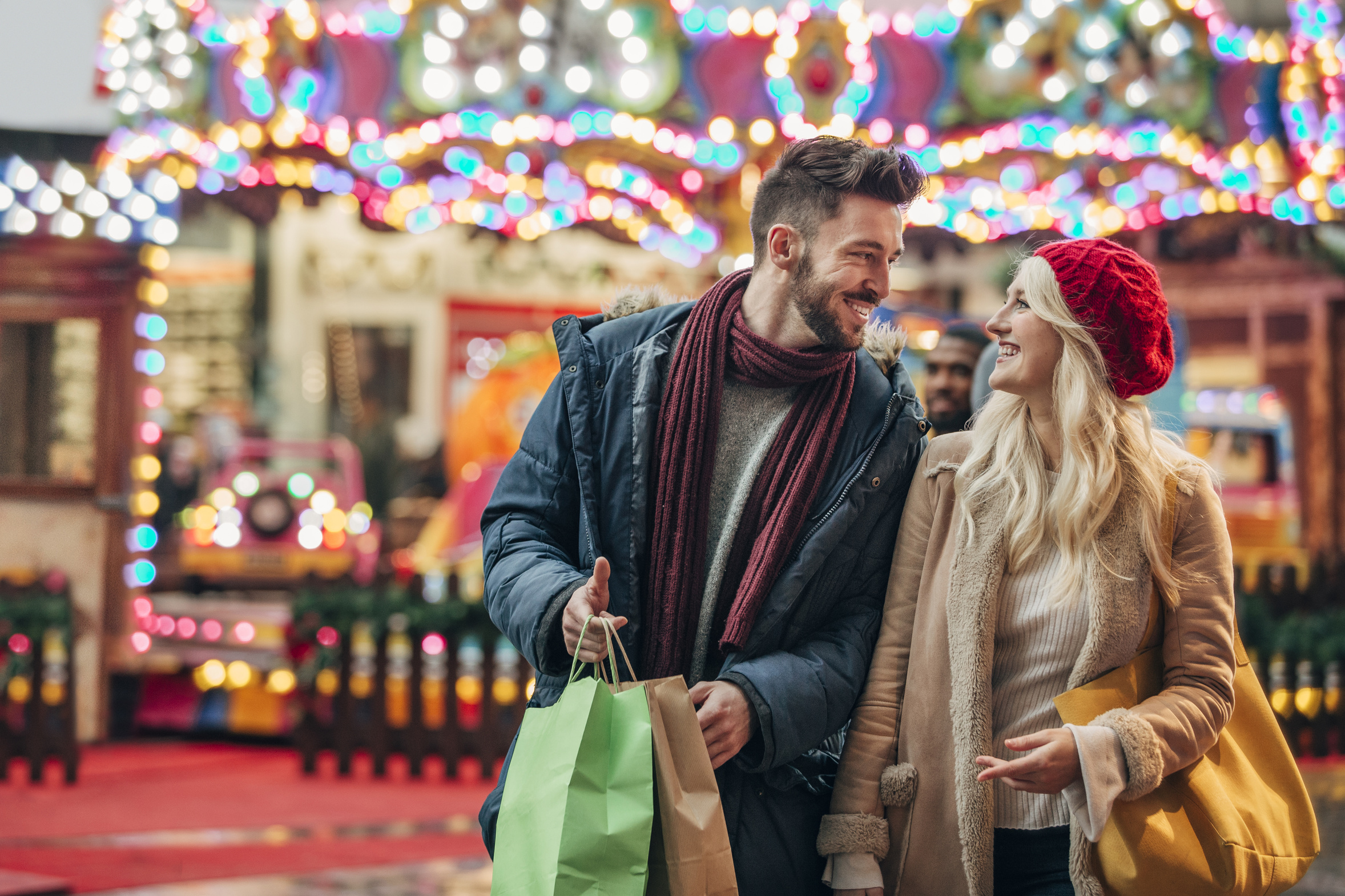 Ein junges Paar am Weihnachtsshoppen in der Stadt, im Hintergrund bunte Lichter eines Karussells