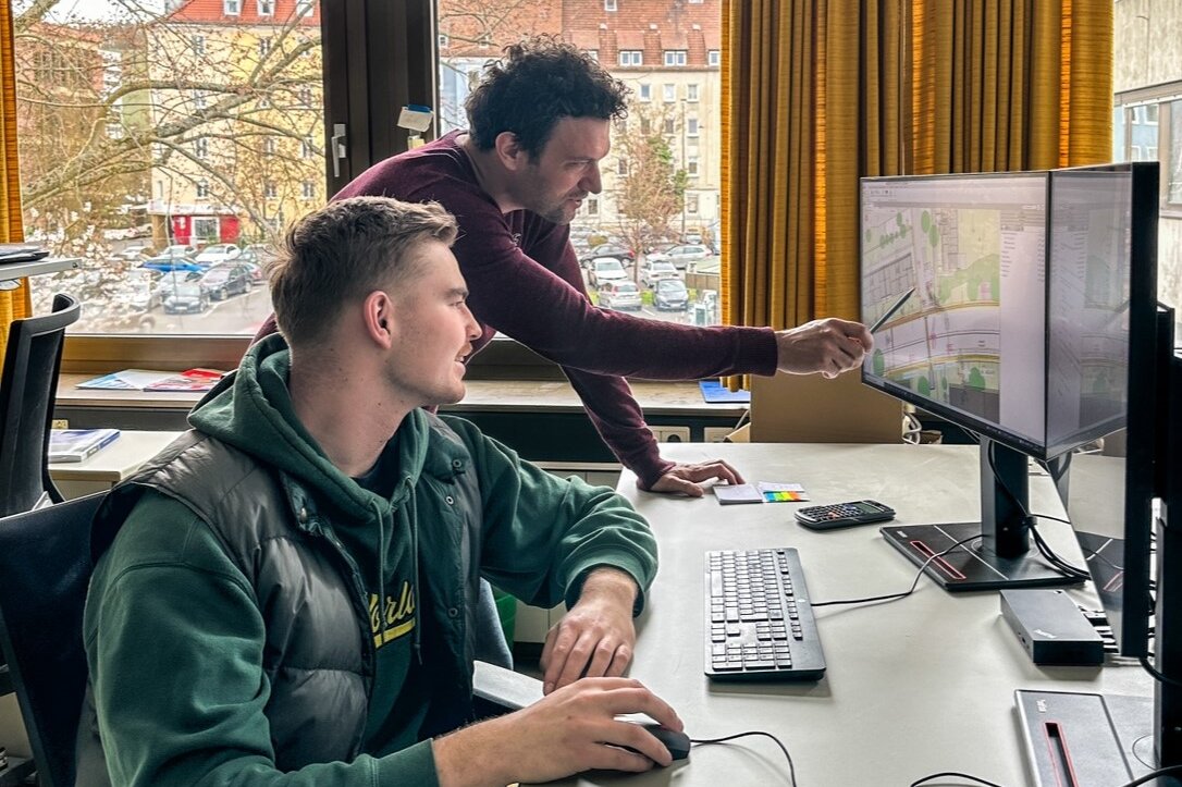 junger Mann hinter PC mit Maus in der Hand, dahinter zeigt der Ausbilder auf den PC