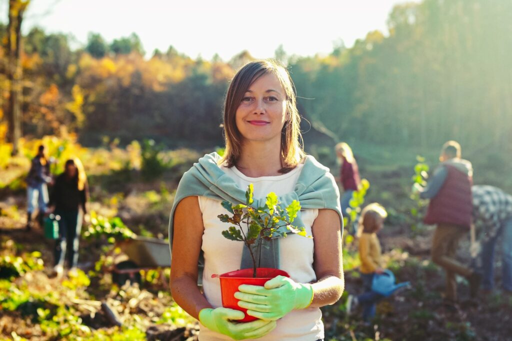 junge Frau hält kleinen Baum in Topf mit Gartenhandschuhen in den Händen und grinst bei Sonnenschein