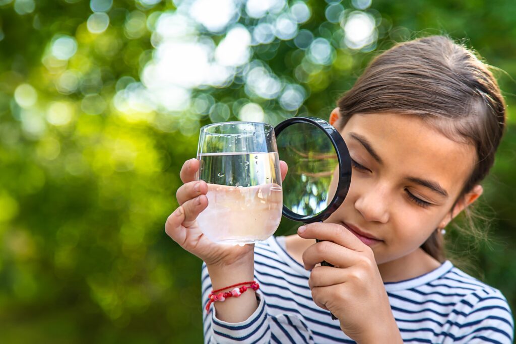 Ein junges Mädchen blickt durch eine Vergrößerungslupe auf ein Wasserglas