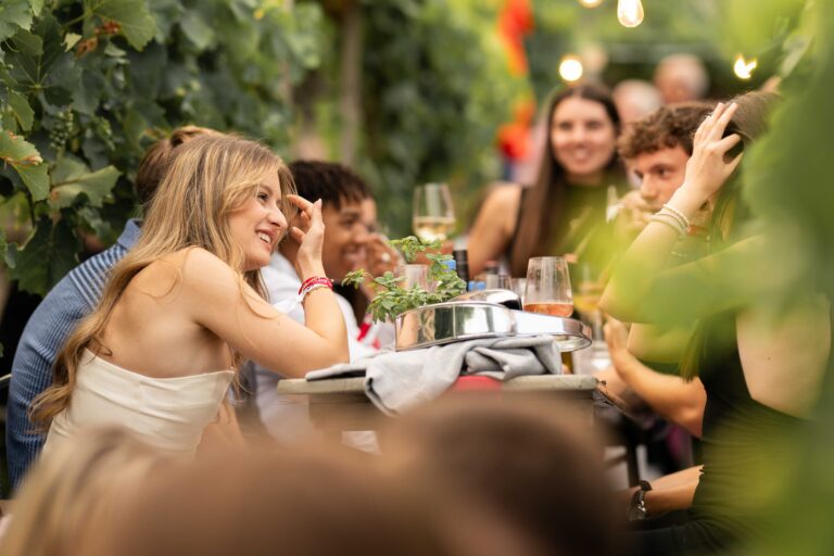 Personen sitzen mit einem Glas Wein an einem Tisch inmitten von Weinreben