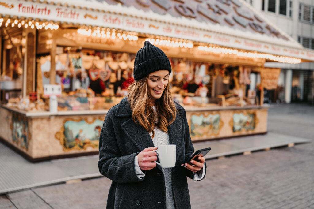 Junge Frau winterlich gekleidet mit Tasse und Handy vor Weihnachtsmarkt-Stand
